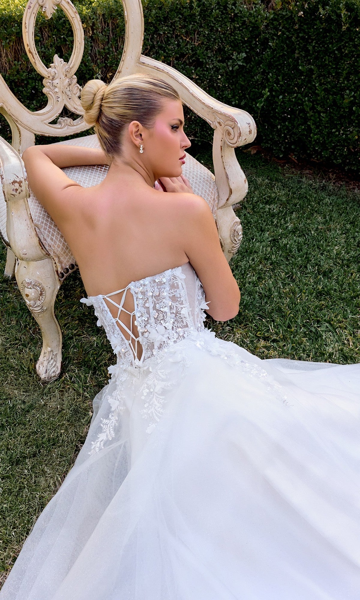 A woman in the Ladivine C174W white formal dress, featuring a strapless corset bodice and lace-up back, sits on grass and leans against an ornate cream-colored chair.